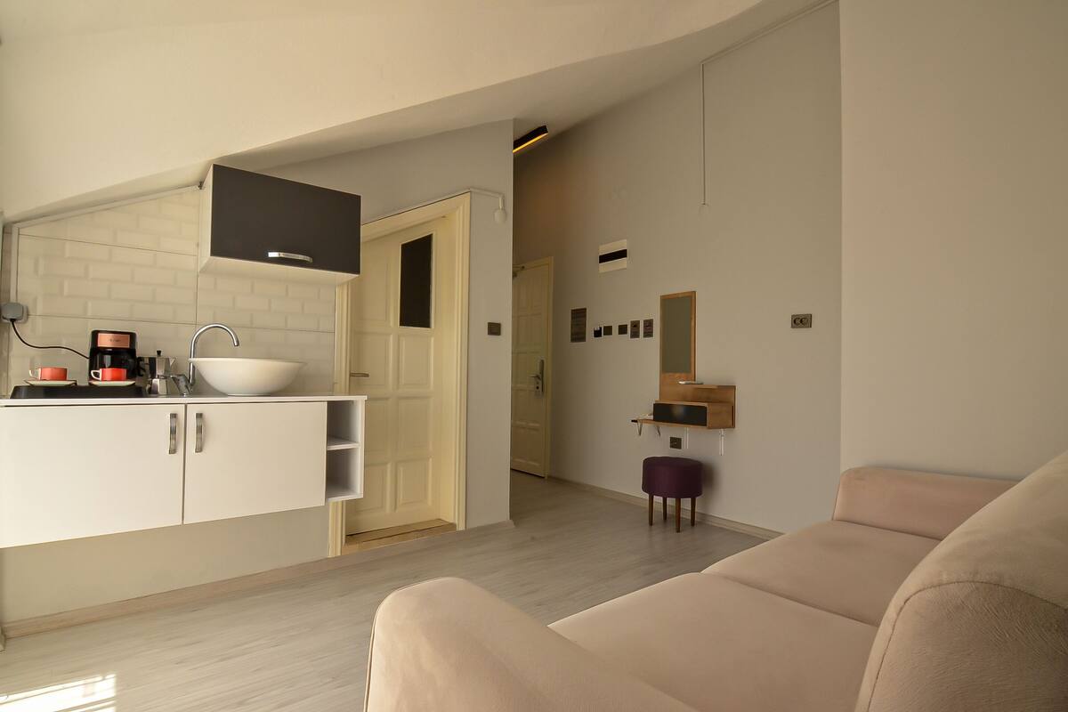 A modern kitchen with a white sink, cabinets, and a coffee maker. There is a beige sofa and a small purple stool in the living area.