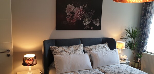 Ein Schlafzimmer mit einem Bett, einem Nachttisch mit Lampe, einer Topfpflanze und einem Blumenbild an der Wand.