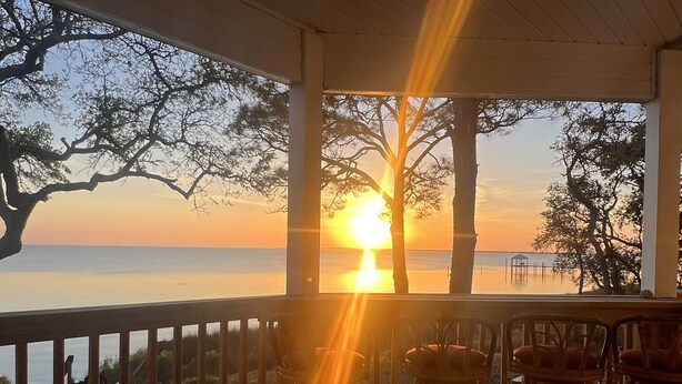 A wooden deck with chairs, overlooking a sunset over the water, framed by trees. - Secluded Waterfront Bay Home! (Eastpoint)