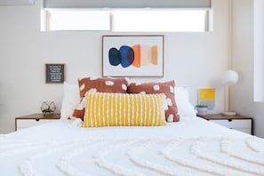 A neatly made bed with white and brown pillows, a framed abstract art piece above, and a small bedside table with a lamp and a plant.