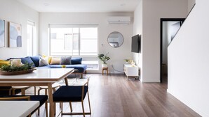 Un salon moderne avec un plancher en bois, un canapé avec des coussins bleus, une table à manger avec des chaises, un téléviseur fixé au mur et une grande fenêtre.