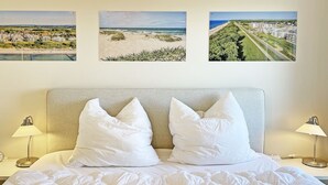 A neatly made bed with white bedding and pillows, two bedside lamps, and three framed pictures on the wall depicting beach and residential scenes. - Deichgraf 07, Direkter Meerblick aus Allen Drei Zimmern, 2 SZ, 2xbad, TG, Kamin (Börgerende-Rethwisch)