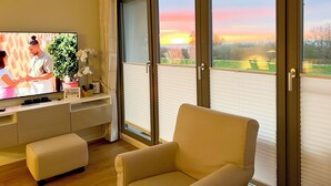 A living room with a television, a sofa, a small table, and a window with blinds. - Deichgraf 07, Direkter Meerblick aus Allen Drei Zimmern, 2 SZ, 2xbad, TG, Kamin (Börgerende-Rethwisch)