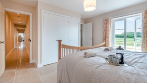 A bedroom with a bed, a bedside table with a kettle and mug, a window with curtains, and a hallway leading to a door.