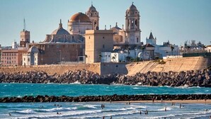 A coastal cityscape with a prominent historical building featuring domes and bell towers, a rocky shoreline, and a beach with people swimming. - Precioso Apartamento EN Planta Baja A Estrenar (MIJAS-COSTA)