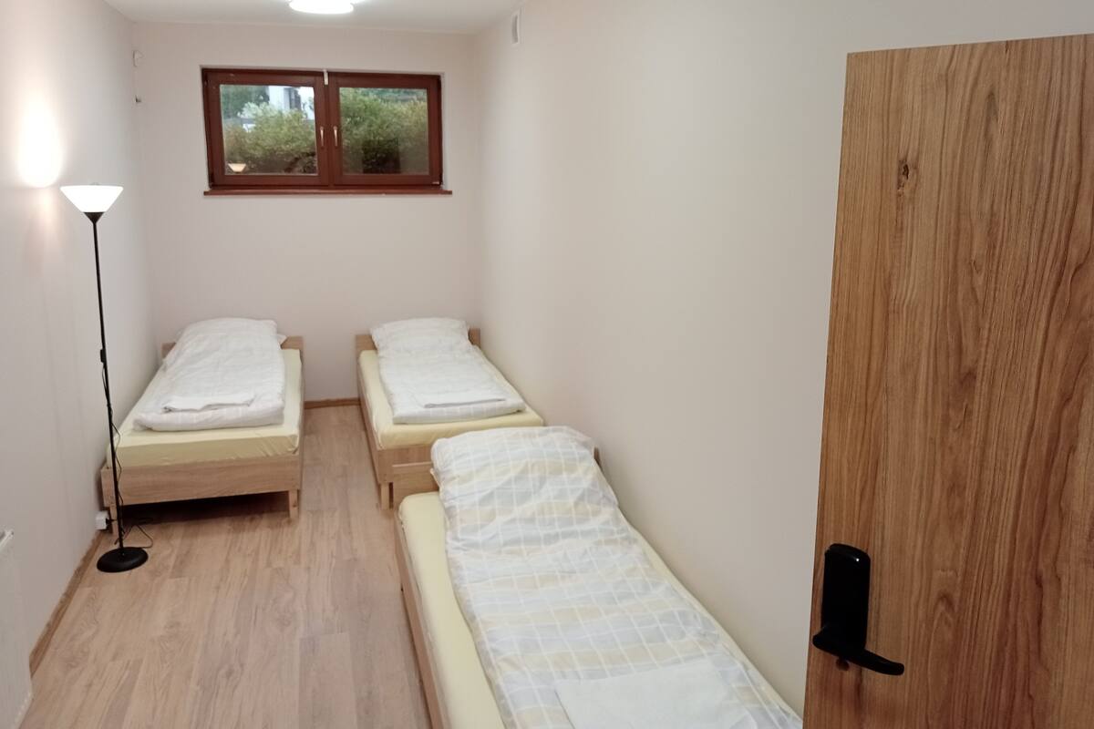 A small, clean room with two single beds, a wooden door, a window with a view of greenery, and a floor lamp.