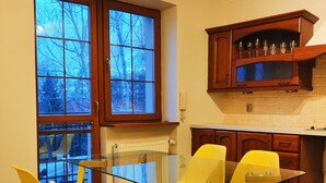 Une cuisine moderne avec une table à manger en verre et des chaises jaunes, un meuble en bois contenant de la verrerie, et une grande fenêtre donnant sur des arbres sans feuilles.
