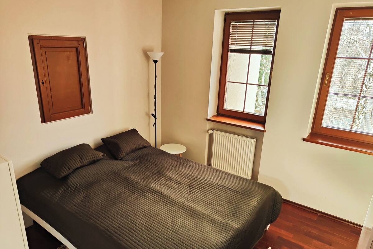 A bedroom with a bed, a radiator, a window with blinds, a floor lamp, and a wooden door.