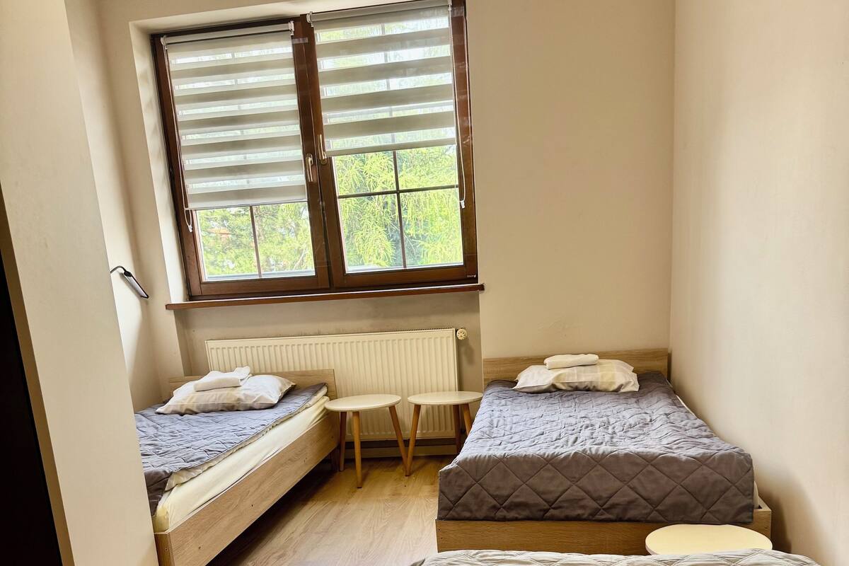 A small, well-lit room with two single beds, a small table, and a window with blinds.