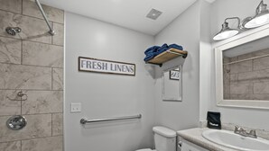 Baño con inodoro, lavamanos y ducha. Hay un cartel que dice "FRESH LINENS" y un perchero con toallas.