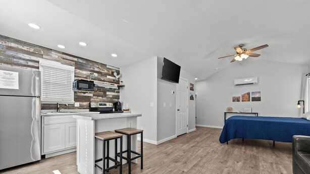 Una cocina moderna con gabinetes blancos, un protector contra salpicaduras de madera y un microondas integrado. Hay una zona de bar con taburetes, un ventilador de techo y una cama con colcha azul en la habitación contigua.