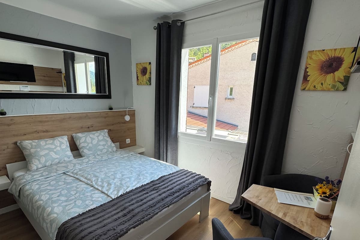 A bedroom with a bed, a desk, a chair, a ceiling fan, and a window with curtains.
