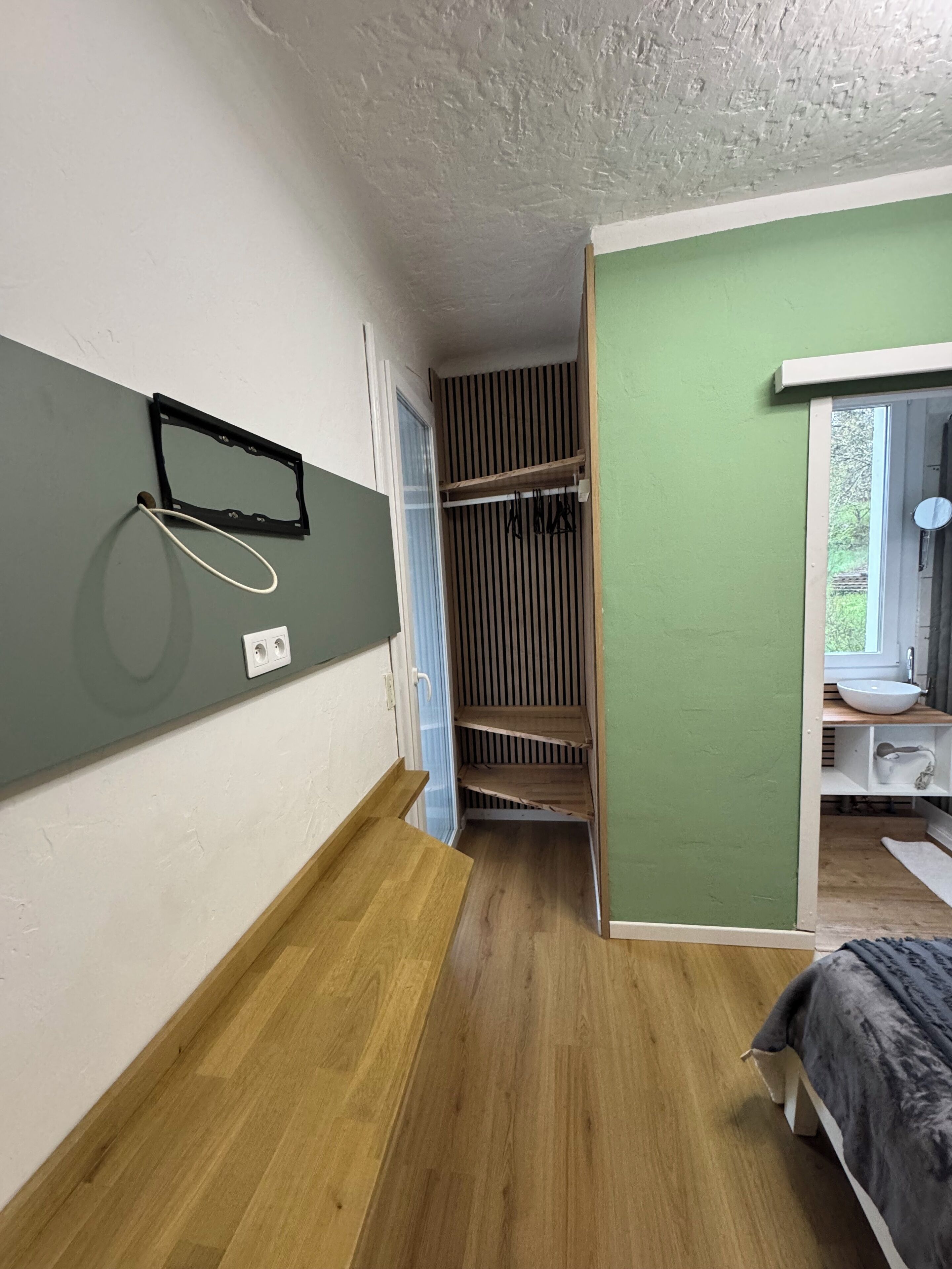 A room with a wooden floor, a green wall, a white ceiling, a window with a view, a bed, a small table, and a mirror on the wall.