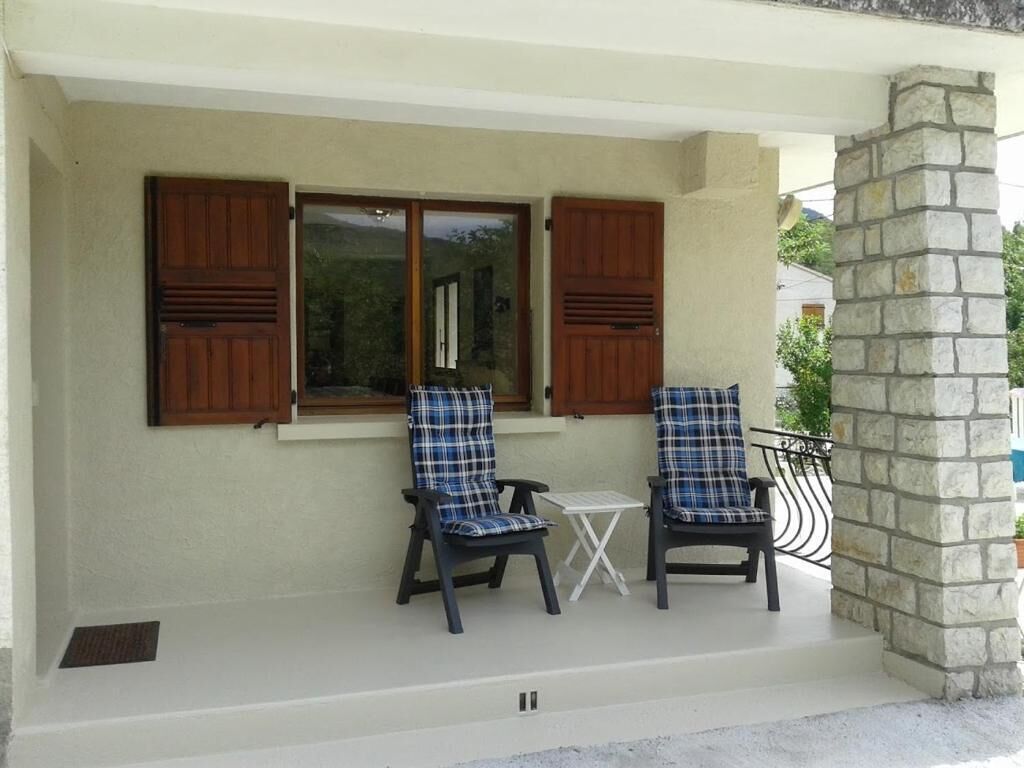 A covered porch with two chairs and a small table, wooden shutters, and a stone pillar.