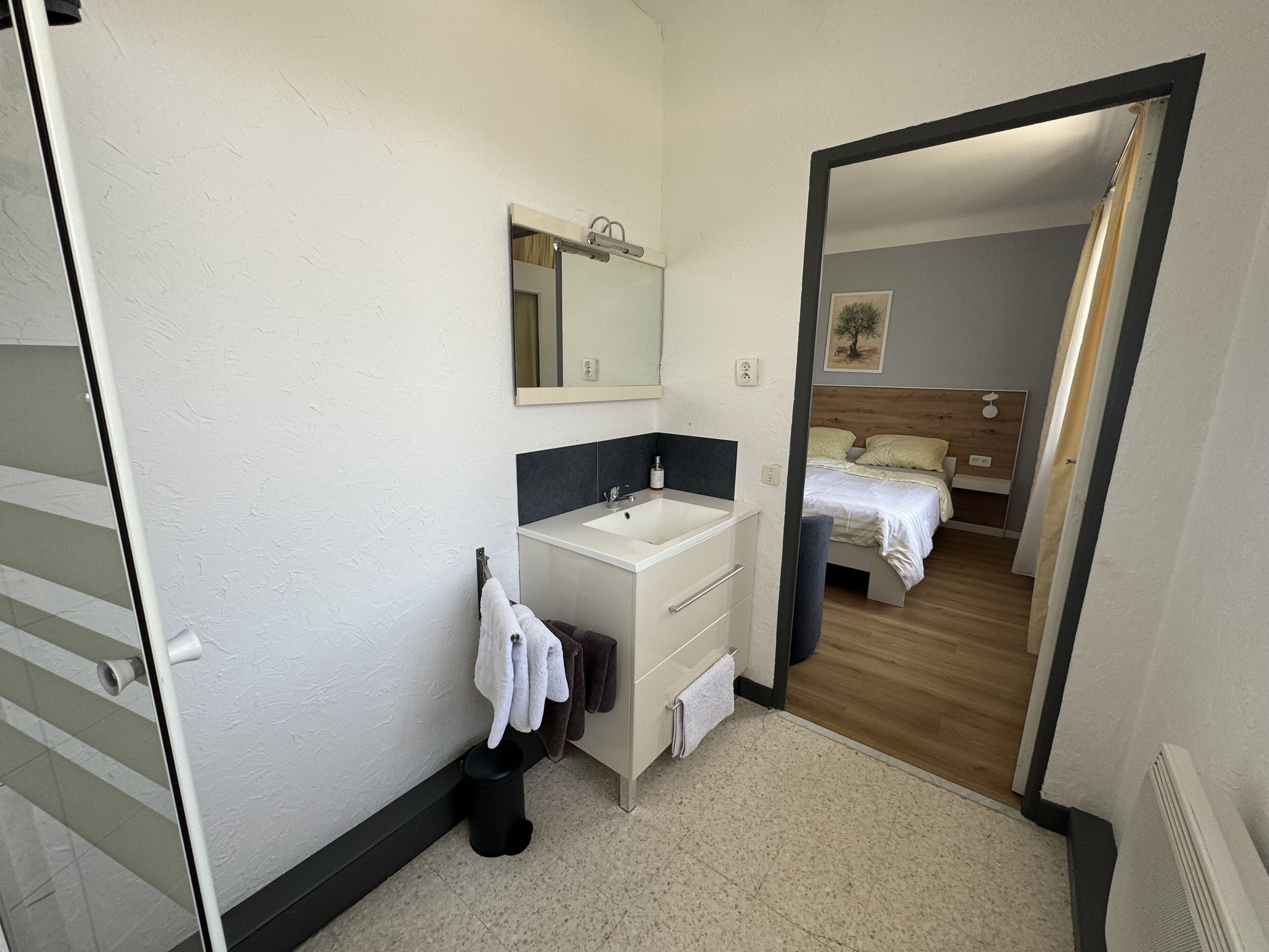 Ein Badezimmer mit weißem Waschbecken, Spiegel und Handtuchhalter. Eine Tür führt zu einem Schlafzimmer mit Bett und Nachttisch.