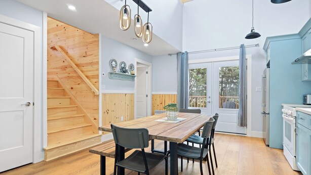 A spacious kitchen with a wooden staircase, a dining area with a wooden table and chairs, and a view of the outdoors through large windows. - Avonlea Forest Hill Cottages (Cavendish)