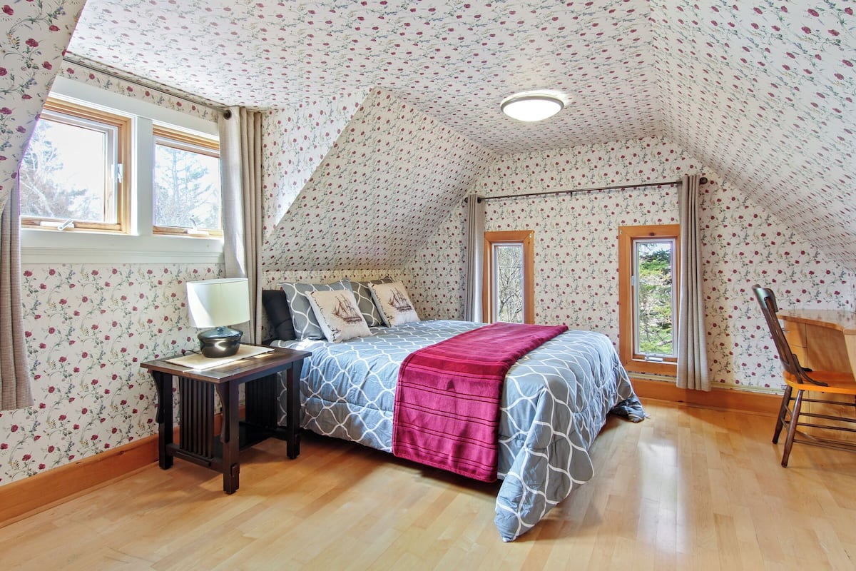 A bedroom with a sloped ceiling, floral wallpaper, a bed with patterned bedding, a bedside table with a lamp, and a window with curtains.