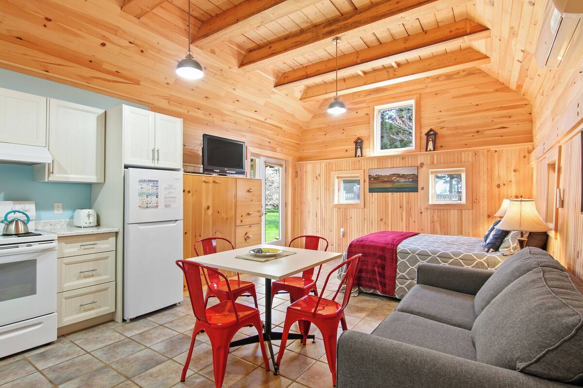 A cozy cabin interior with wooden walls, a dining area with red chairs, a kitchenette, a sofa, and a television.