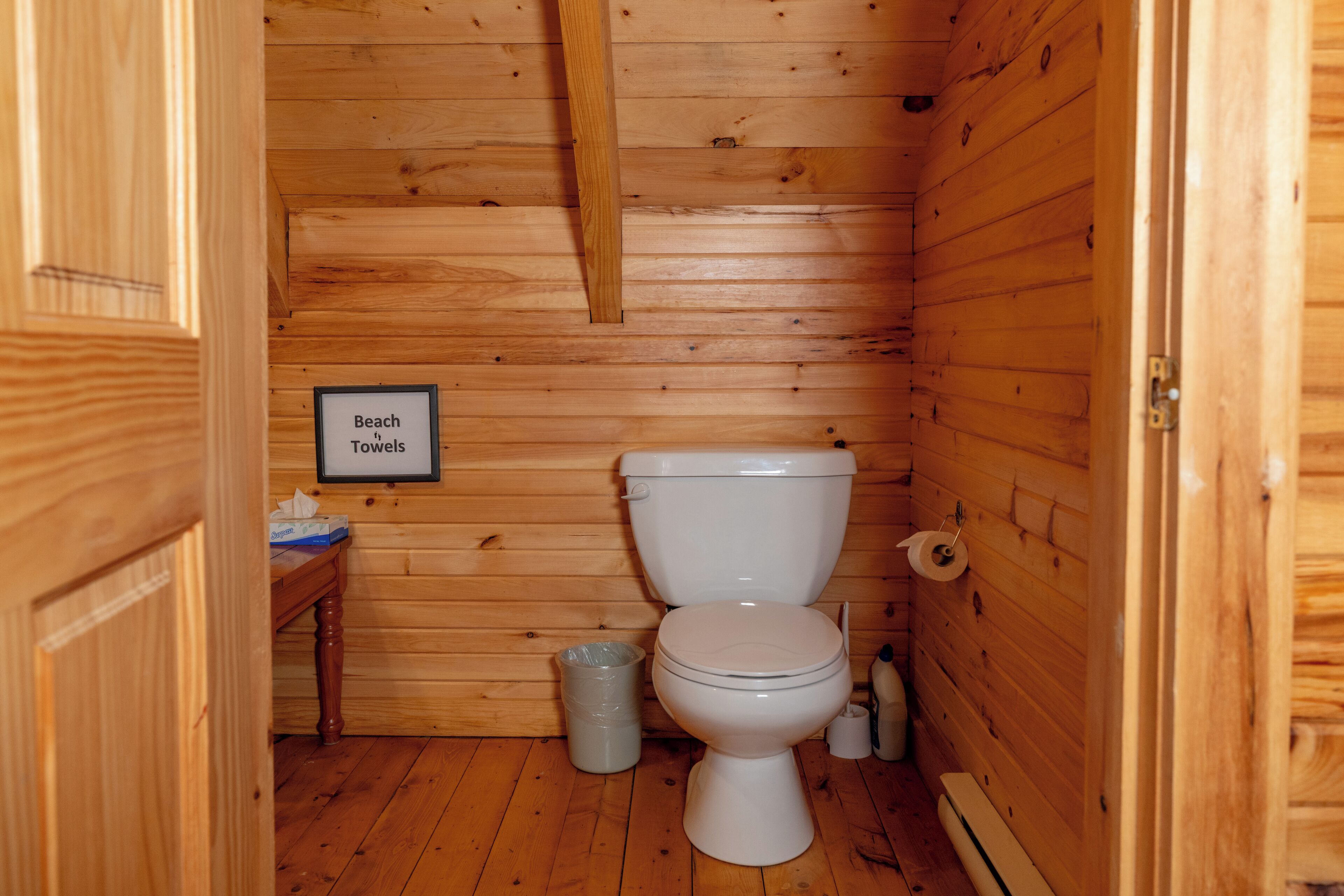 A wooden cabin bathroom with a toilet, a trash can, and a sign that reads 'Beach Towels'.