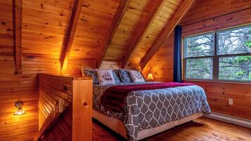 Quarto de cabana de madeira com uma cama, almofadas, janela e mesa de cabeceira.