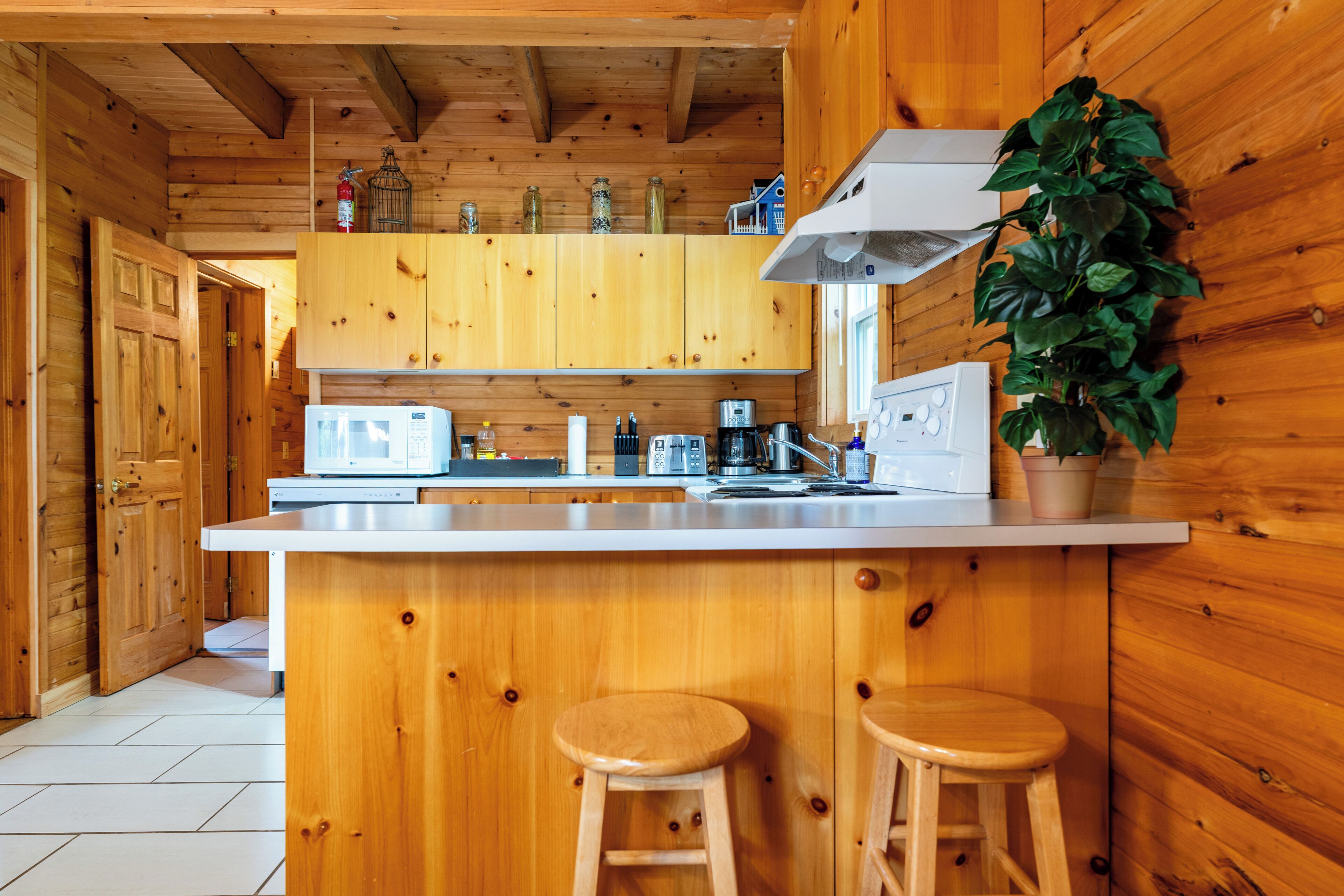 A wooden cabin interior with a kitchen area featuring a white countertop, wooden stools, a stove, and a microwave.