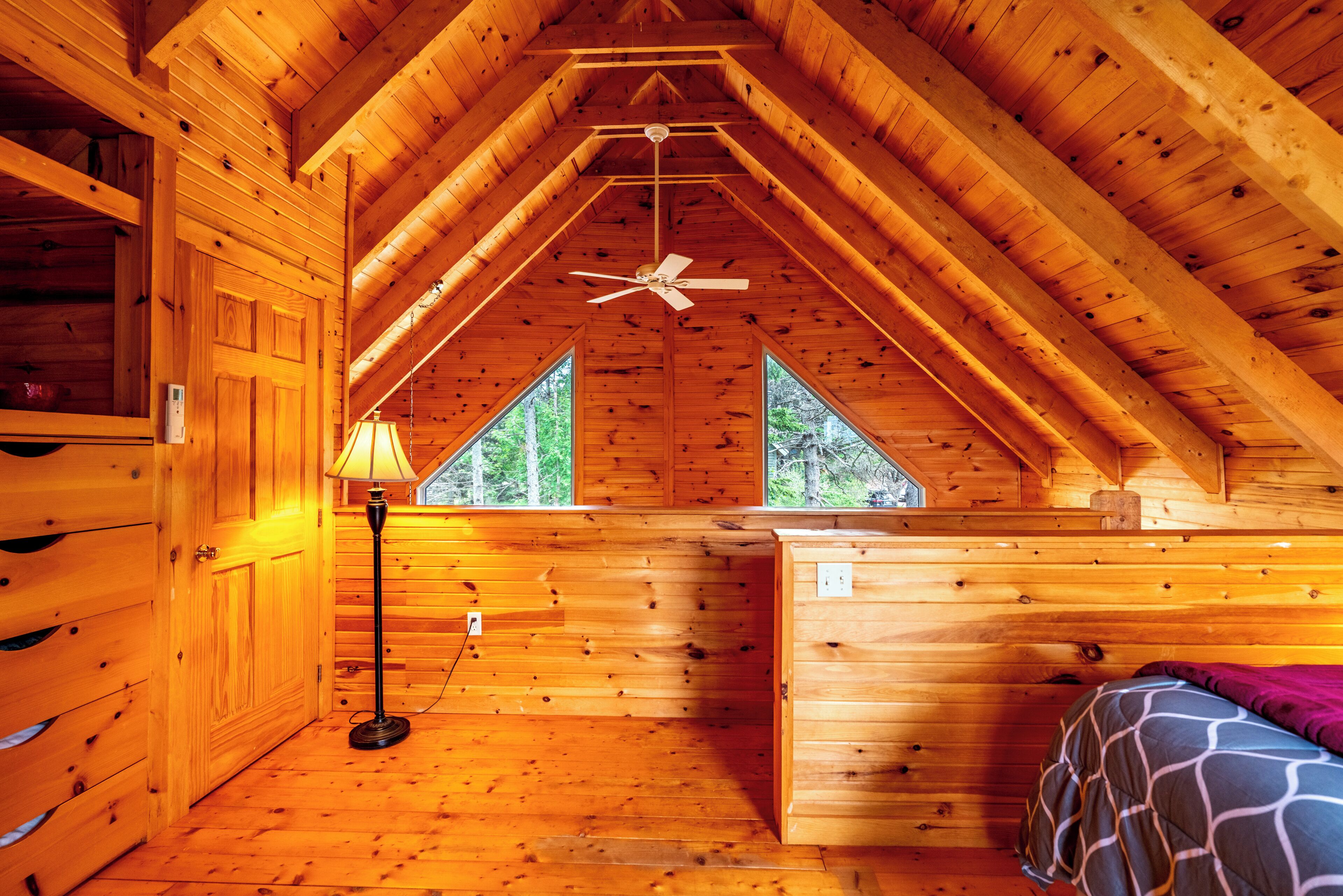 A cabin with a sloped ceiling, a ceiling fan, a bed, a desk, and a window with a view of trees.