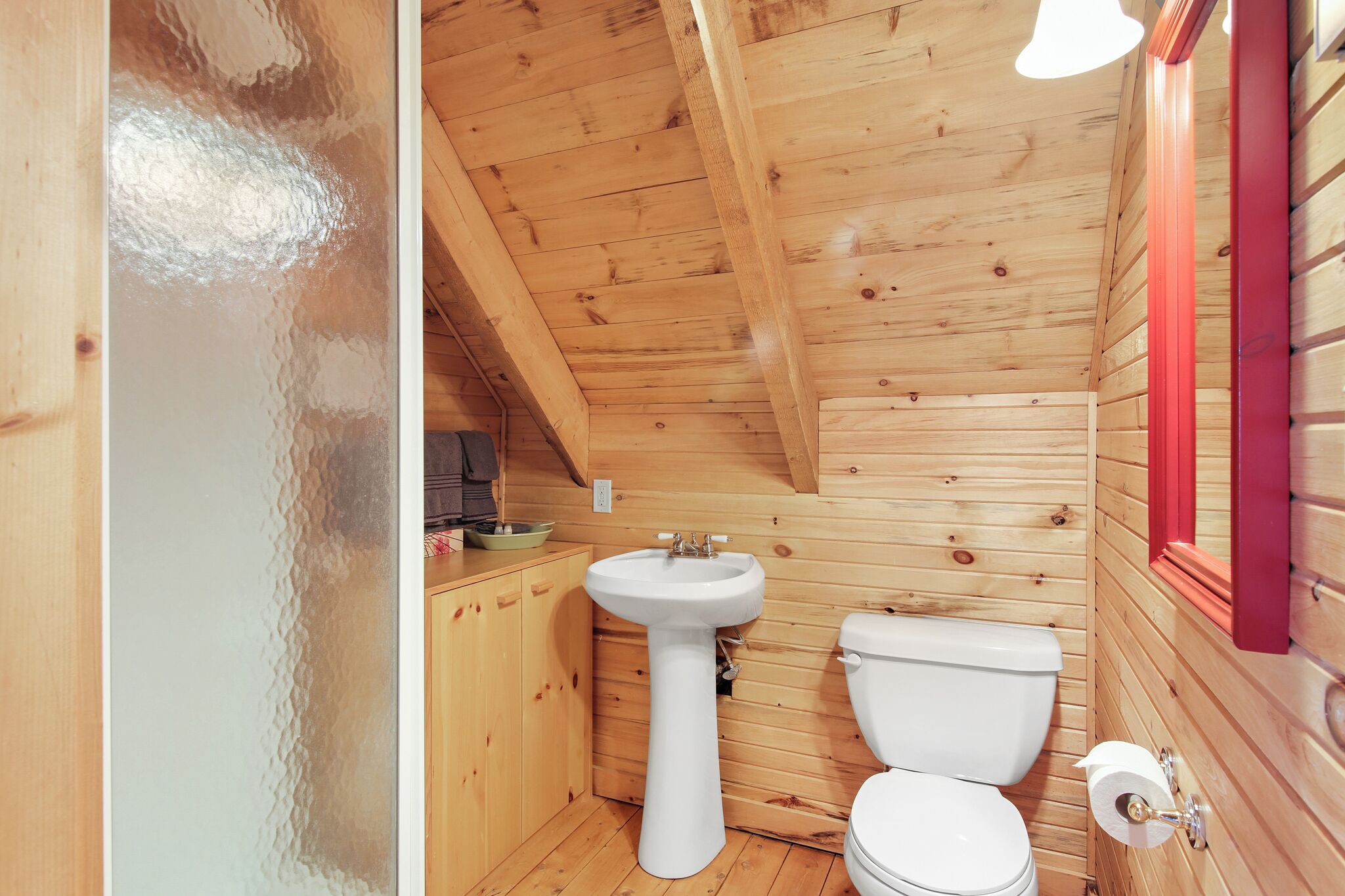 A small, wooden cabin bathroom with a white sink, toilet, and wooden cabinets.
