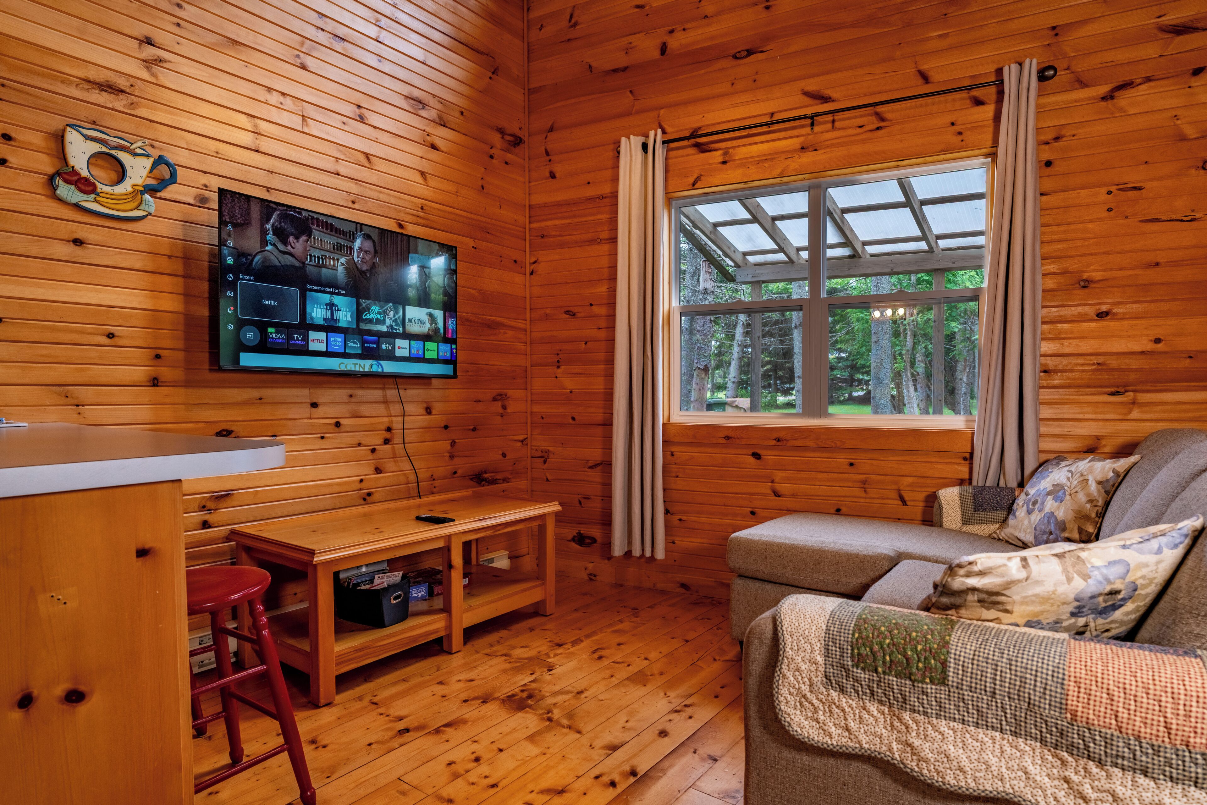 Un intérieur en bois avec un téléviseur fixé au mur, un canapé, une petite table et une fenêtre avec des rideaux.