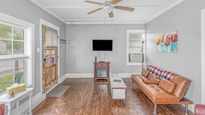 A living room with a wooden floor, a ceiling fan, a TV, a couch, and a painting on the wall. - Close to Hospital and Downtown, Eastside Bungalow, Dogs Welcome + Fee (Florence)