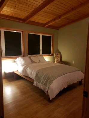 Un dormitorio con techo de madera, una cama grande con ropa blanca y dos ventanas con persianas.