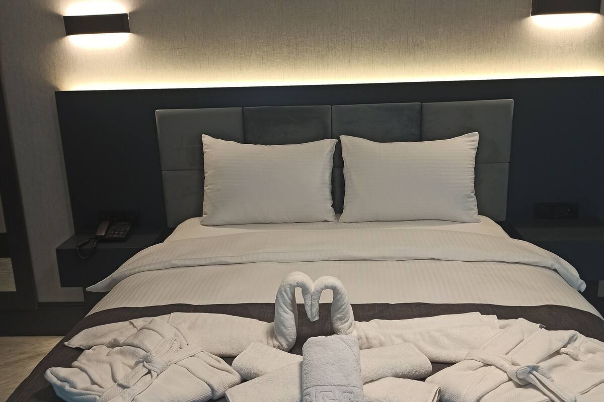 Una cama bien tendida con sábanas blancas, un arreglo de toallas en forma de corazón y dos lámparas de noche.