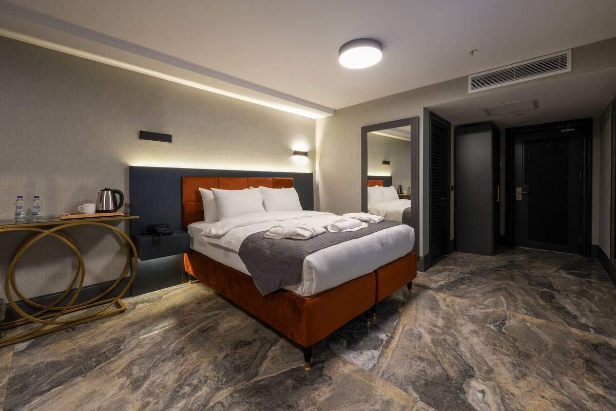 Una habitación de hotel moderna con suelo de mármol, una cama grande con cabecero, mesitas de noche, un espejo y una puerta.