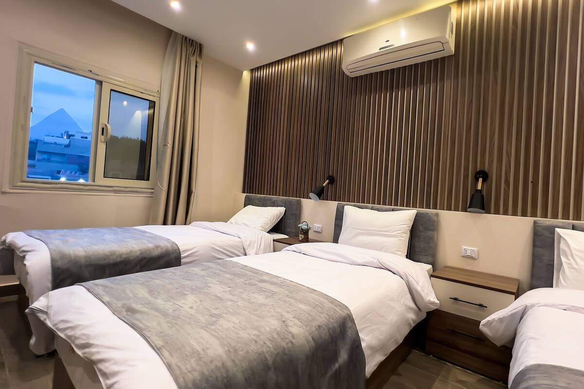 A hotel room with two beds, a window with a view of a mountain, an air conditioning unit, and a wall-mounted lamp.