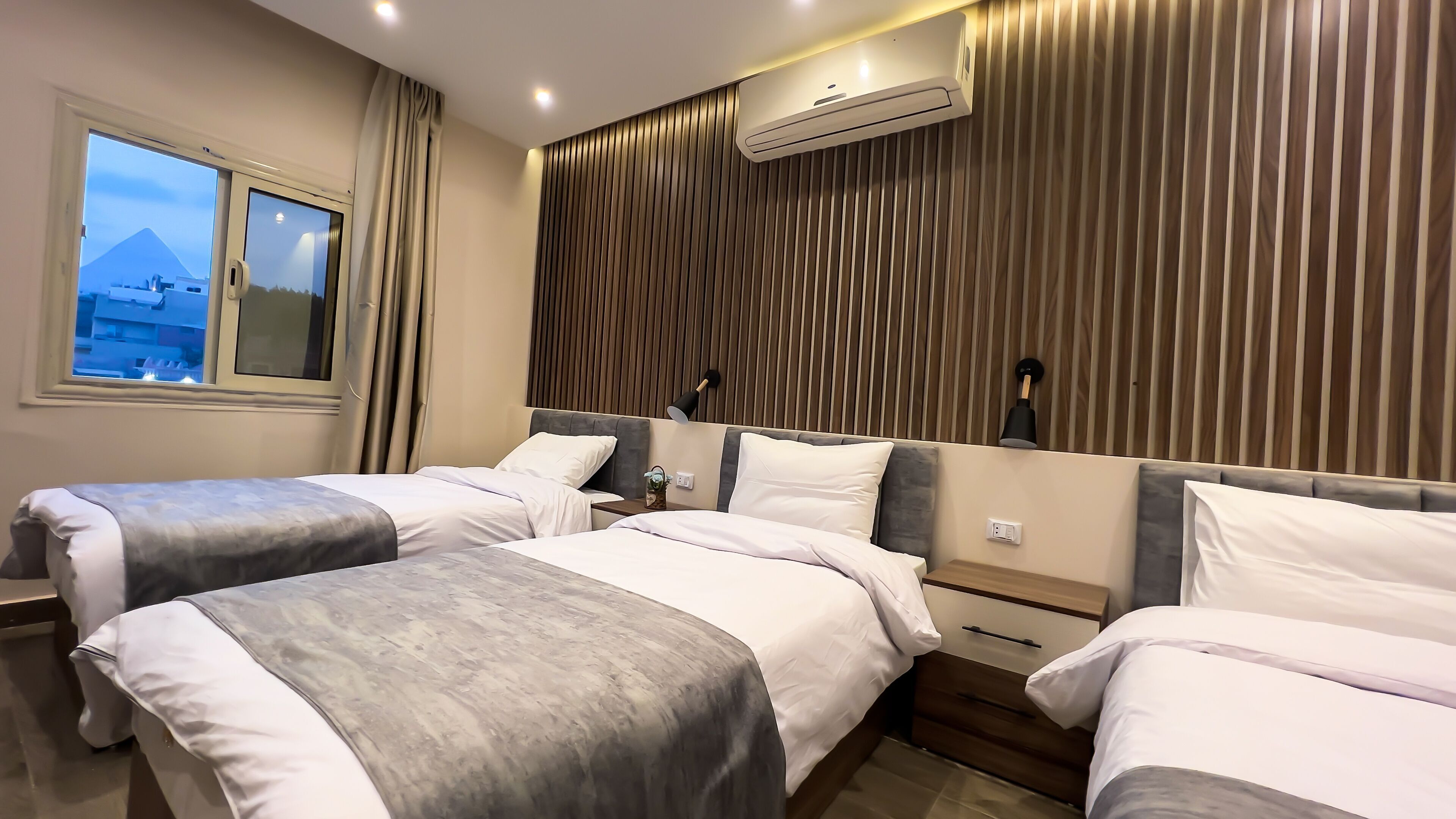 Habitación de hotel con dos camas, una ventana que da a edificios, un equipo de aire acondicionado y una lámpara fijada a la pared.