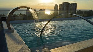 Una piscina con un elemento acuático, rodeada por una terraza con azulejos y vista a un paisaje urbano.