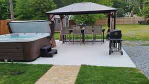 Un patio trasero con jacuzzi, mesa y sillas en un porche cubierto, y una parrilla.