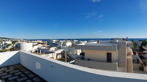 Um terraço no telhado com vista para o mar, edifícios brancos e céu azul claro.