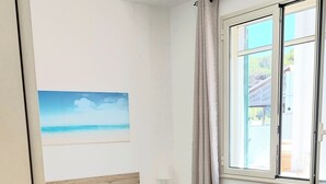 Uma cama de solteiro com colcha verde e toalhas brancas, mesa de cabeceira com abajur, janela com cortinas e um quadro de uma praia na parede.