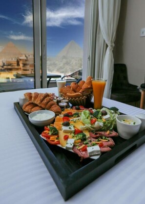 Bandeja de desayuno con croissants, queso, embutidos y verduras, con vistas a una pirámide al fondo.