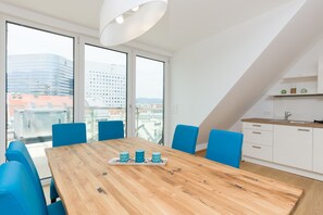 A modern room with a wooden table and blue chairs, large windows with a city view, and a kitchen area.