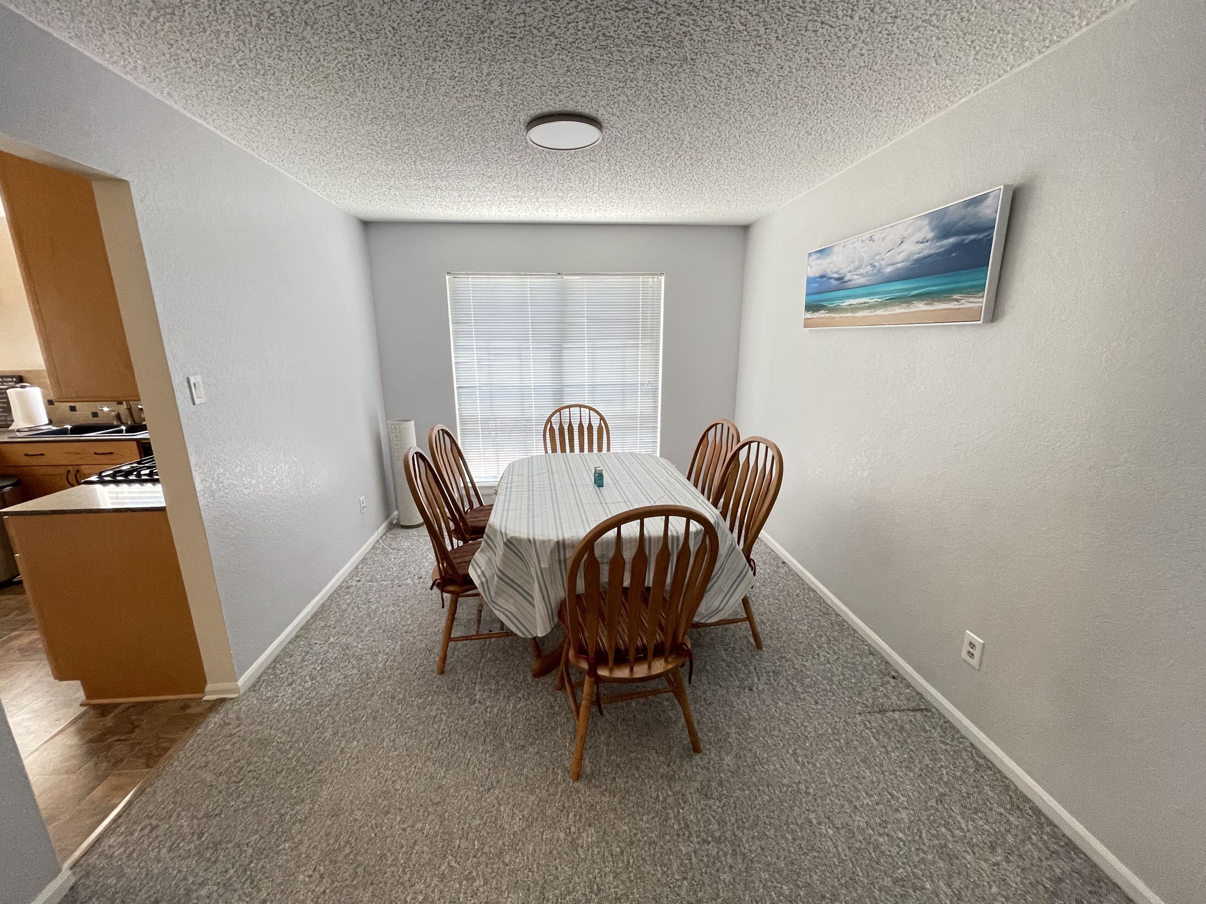 A dining area with a round table and six wooden chairs, a window with blinds, and a painting on the wall.