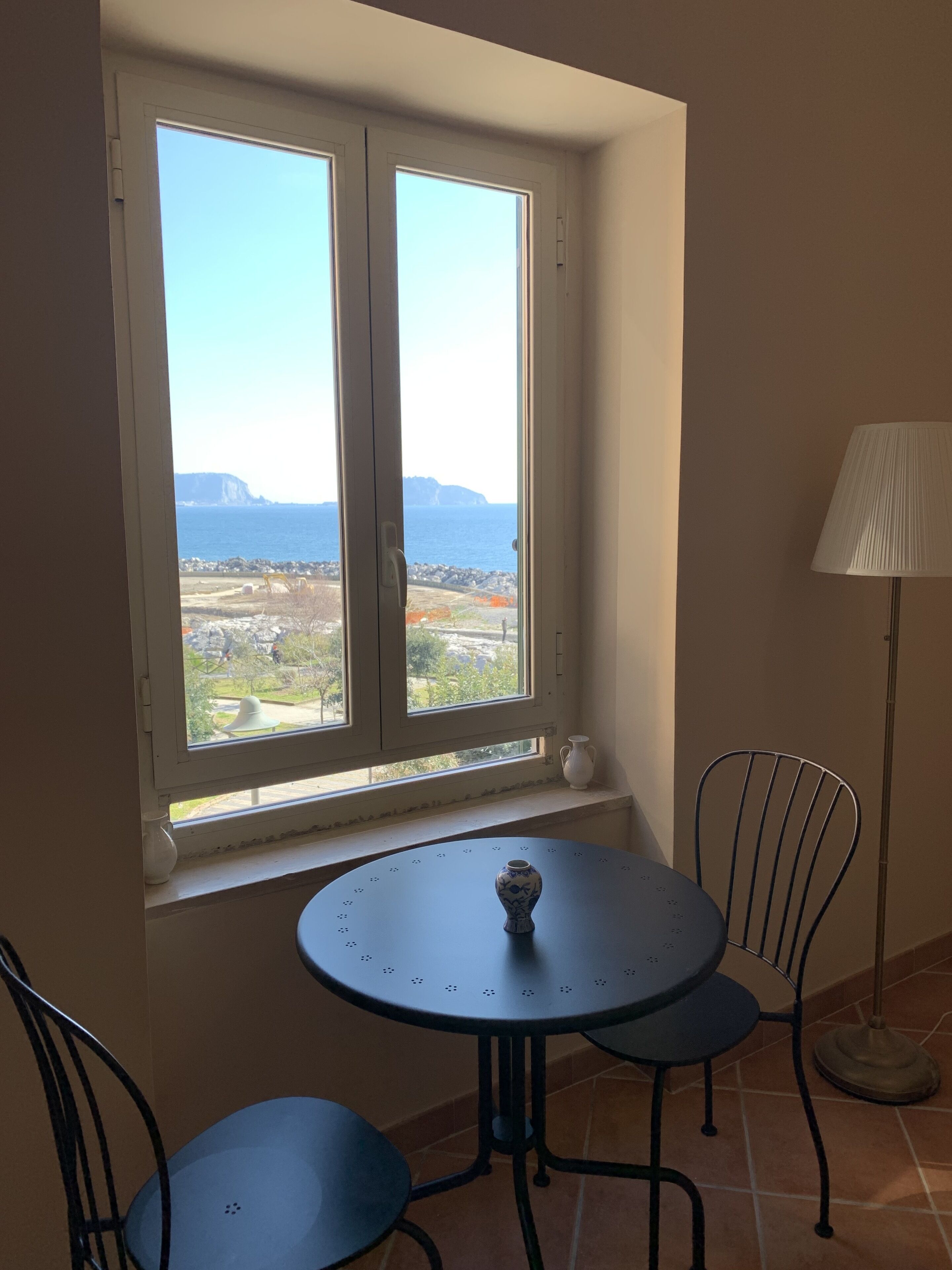 A room with a window overlooking a coastal landscape, a round table with two chairs, and a floor lamp.