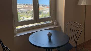 A room with a window overlooking a coastal landscape, a round table with two chairs, and a floor lamp.