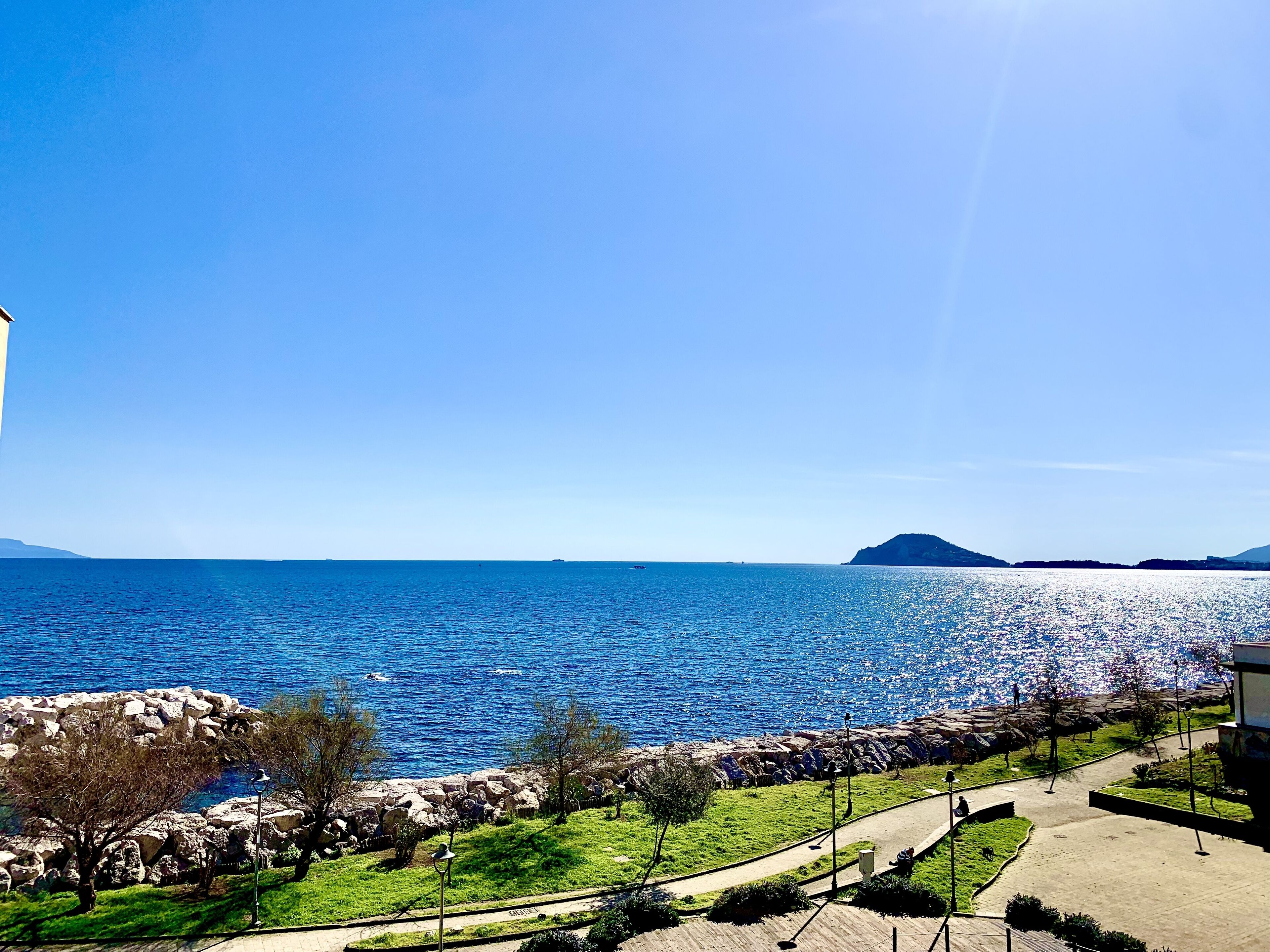 Vista costera con cielo despejado, costa rocosa y un paseo pavimentado a orillas del agua.