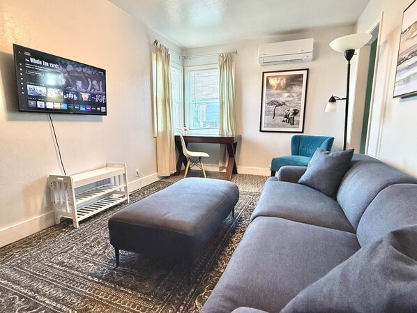 A living room with a flat-screen TV, a sofa, an ottoman, a desk, and a chair.
