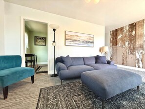 A living room with a sectional sofa, a teal armchair, a patterned rug, a floor lamp, and a large abstract painting.