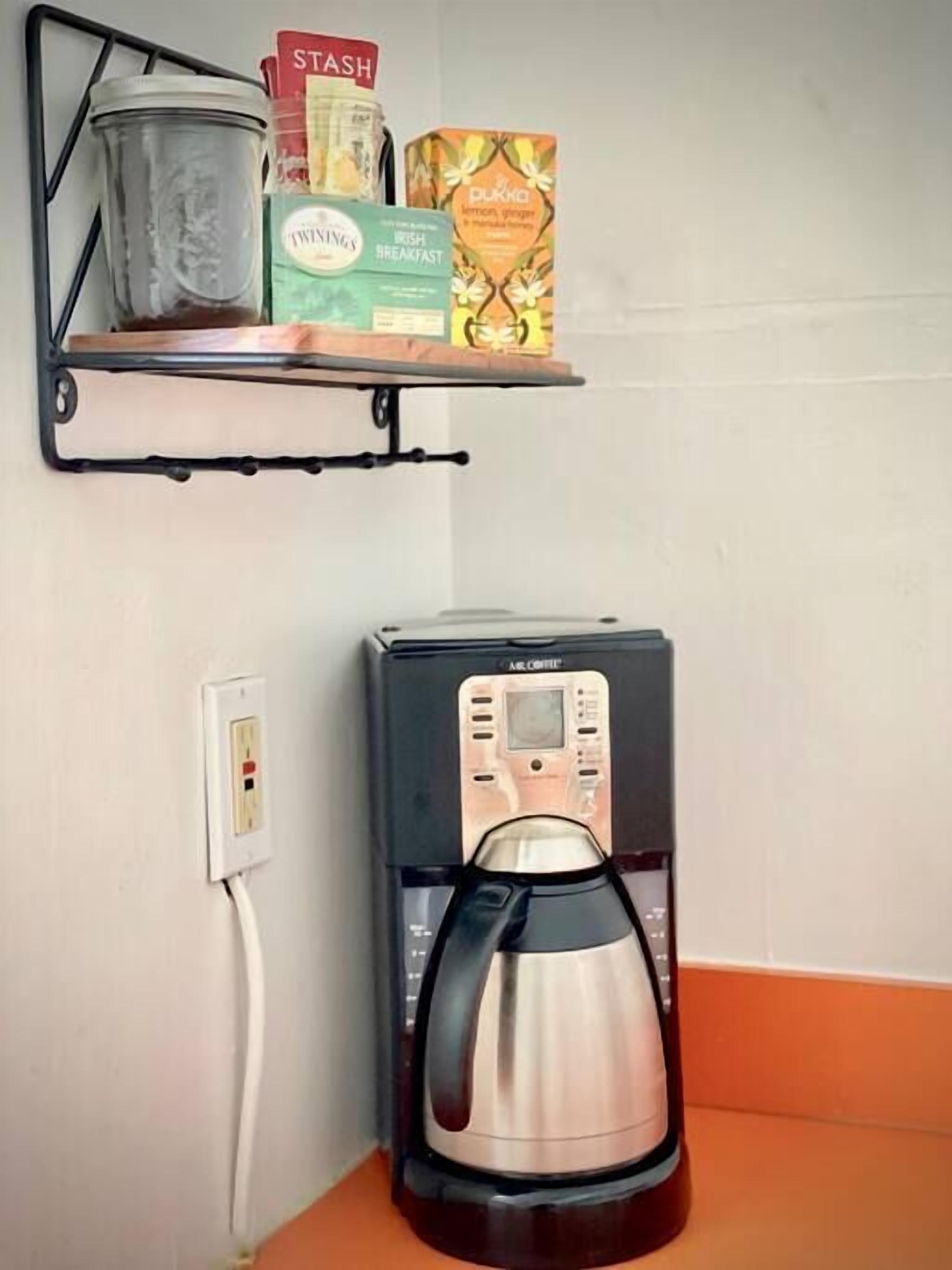 Une machine à café est posée sur un plan de travail, et une étagère au-dessus abrite une poubelle, une boîte de café Twinings et une boîte de thé.