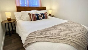 A neatly made bed with a white comforter and a beige knitted throw, two bedside lamps, and a window with blinds. - Central Craftsman Apartment (Arcata)
