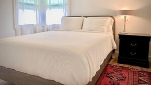 A neatly made bed with a white duvet, a bedside table with a lamp, and a red patterned rug.