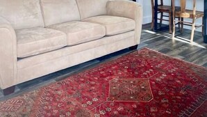 A living room with a beige sofa, a red patterned rug, and a dining area with wooden chairs and a table.
